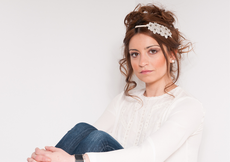 Retrato de moda a Sharai que está sentada con las manos entrelazadas en las piernas  y viste un pantalón vaquero con camisola blanca y un tocado en el pelo realizado por Leyre Benito. Fotografía realizada por Alberto Cossío Fotografía de moda